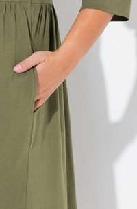 Vestido verde oliva con mangas cortas, que presenta un bolsillo lateral. Tejido suave y ligero con un detalle de cintura fruncida.