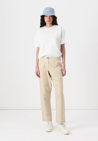T-shirt bianca in cotone con testo giallo, pantaloni beige in cotone e cappello a secchiello blu chiaro. Sneakers bianche con suole gialle.