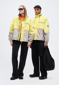 Δύο μοντέλα φοράνε κίτρινες και μπεζ μπουφάν Tommy Jeans, μαύρο παντελόνι, μαύρα παπούτσια· το ένα κρατά μαύρη τσάντα, το άλλο φορά γυαλιά ηλίου.