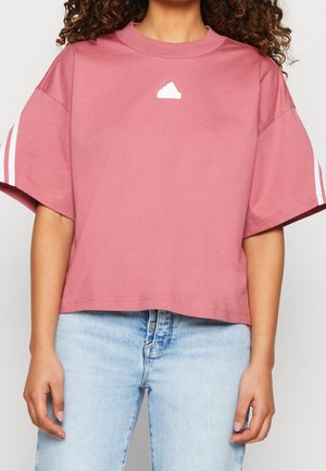 Kobieta nosząca luźny różowy krótki t-shirt z białymi paskami na rękawach i białym geometrycznym logo na piersi, w połączeniu z jasnoniebieskimi dżinsami.