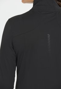 Zwarte jas met hoge kraag, gladde textuur en verticale streepdetails op de achterkant. Ontworpen voor comfort en bewegingsvrijheid.