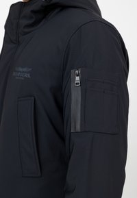 Schwarze wasserfeste Jacke mit einer Reißverschlusstasche am linken Arm, doppeltem Taschen-Design auf der Vorderseite und einem Logo mit Flügeldetail.