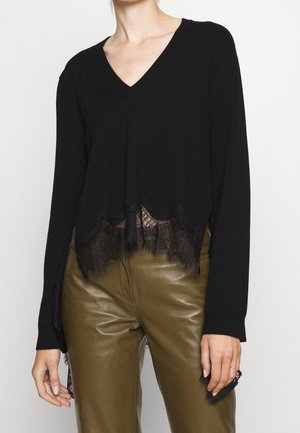 Femme portant un haut noir à manches longues avec col en V et bordure en dentelle, et un pantalon en cuir vert olive, vue du cou jusqu'aux hanches.