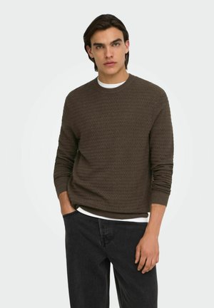 Homme aux cheveux foncés portant un pull marron texturé par-dessus une chemise blanche et un jean noir, debout avec une main dans la poche devant un fond blanc.