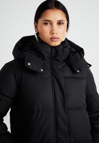 Chaqueta acolchada negra con un alto cuello, botones de presión y una capucha. Presenta paneles acolchados y un acabado de tela lisa y texturizada.
