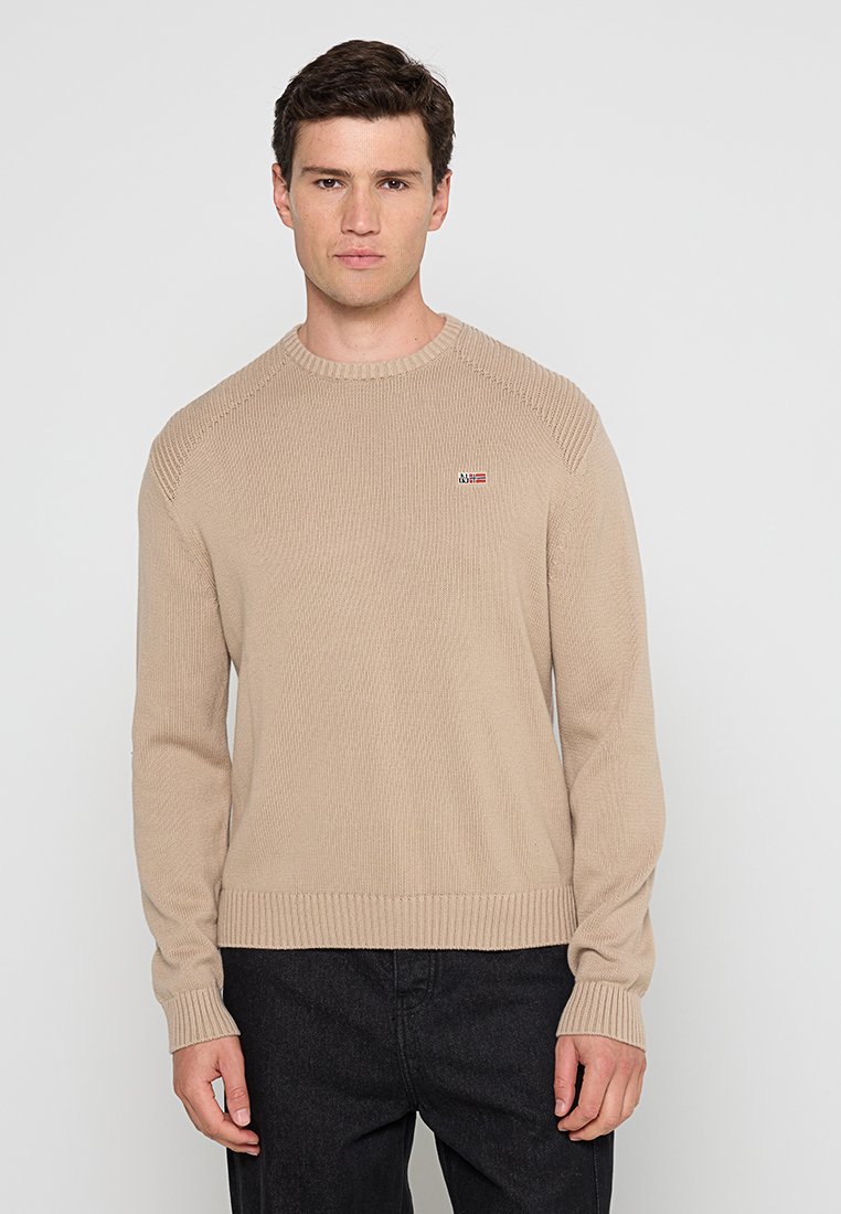 Mężczyzna w beżowym swetrze dzianym z małym logo na piersi i czarnych spodniach, stojący na jasnym jednolitym tle.