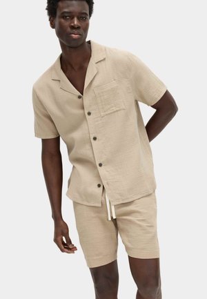 Junger Mann trägt ein beige kurzärmeliges Hemd mit Knöpfen, Brusttasche und passenden Shorts vor weißem Hintergrund.