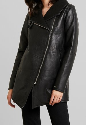 Kobieta w czarnej, asymetrycznej, skórzanej kurtce na zamek z szerokim kołnierzem i długimi rękawami, w połączeniu z czarnymi spodniami.