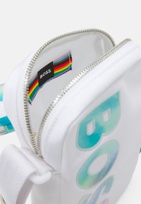 Borsa in tessuto bianco con testo olografico "BOSS", zip argento, etichetta interna a strisce arcobaleno. Strap regolabile in gradiente blu.