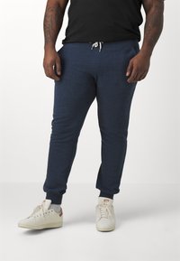 Marineblå joggebukser med elastiske mansjetter, snøring i livet, sidelommer og et teksturert stoff. Båret med hvite sneakers med brune detaljer.