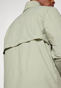 Homme portant une veste vert clair à manches longues avec un détail au dos, vu de dos et légèrement tourné sur le côté.