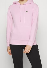Sudadera con capucha de color rosa claro hecha de tela suave que cuenta con un bolsillo frontal, capucha con cordón y un pequeño logo verde. La modelo lleva pantalones negros.
