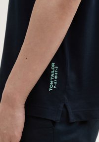 Námořnická modrá tričko vyrobené z bavlny, s rovný střihem, bočními rozparky a mátově zeleným potiskem na dolním pravém rohu.