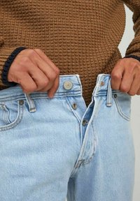 Manos desabrochando unos jeans de mezclilla azul claro que se llevan sobre un suéter de punto marrón, centrándose en el área de la cintura.