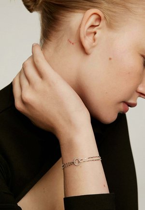Pulsera de plata con un acento circular y pequeños adornos. La mano del modelo reposa contra su cuello, mostrando la joyería.