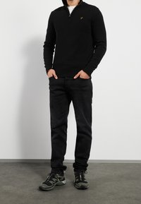 Pull noir à col zippé avec poignets côtelés, porté avec un jean foncé et des baskets noires avec des accents verts. Design simple, sans motifs.