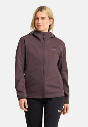 Vrouw draagt een kastanjebruine zip-up hoodie en zwarte broek, staand tegen een effen lichte achtergrond.
