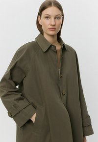 Olíva zöld, hosszú ujjú kabát gombos elülső résszel, nagy gallérral és oldalsó zsebekkel. Tapintása texturált, és laza szabású.