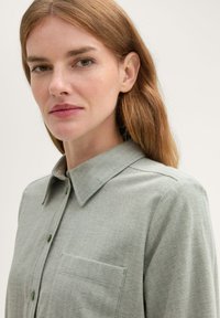 Světle zelená košile s vzorem rybí kosti, se strukturovaným límcem, předními knoflíky a jednou náprsní kapsou. Bavlněný materiál, hladká textura.