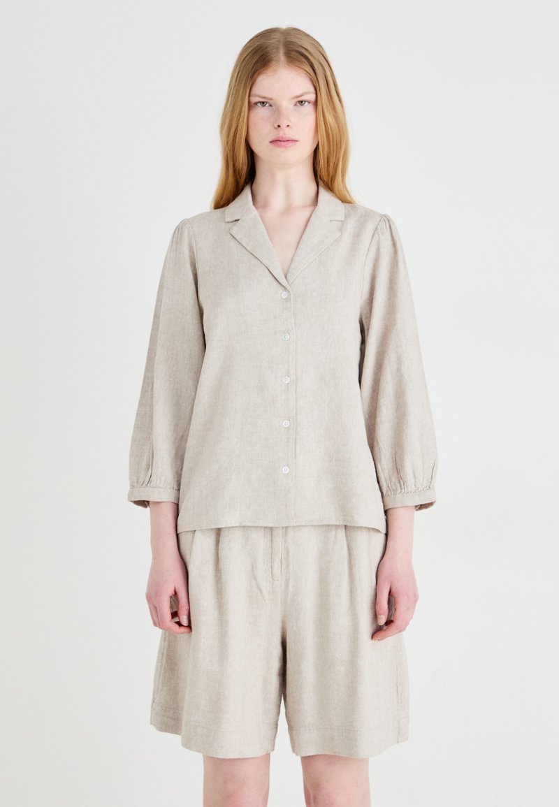 Blusa in lino beige con bottone frontale, colletto a scollo a V e maniche a palloncino, abbinata a pantaloni corti larghi coordinati.
