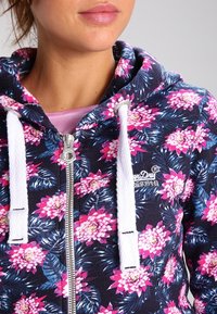 Felpa con zip floreale che presenta uno sfondo blu scuro con fiori rosa e foglie verdi. Stringhe bianche e zip in metallo argentato.
