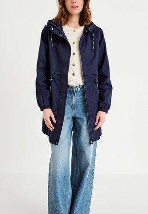 Woman wearing navy hooded rain jacket over white buttoned sweater and wide-leg blue jeans, standing against plain background.