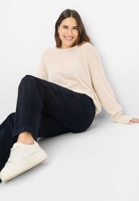 Maglione beige, leggero, con una vestibilità ampia, abbinato a pantaloni larghi blu scuro. Il modello indossa sneaker bianche con suola spessa.