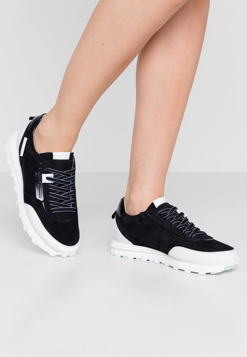 Baskets en daim noir avec semelles en caoutchouc blanc, dotées d'une tige texturée, de lacets noirs et d'un accent brillant noir au talon.