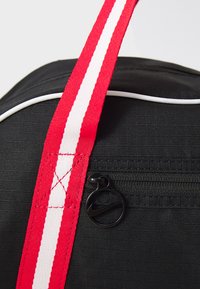 Black backpack with a textured finish, featuring a wide red strap with white stripes, and a black zipper pull with a circular logo.