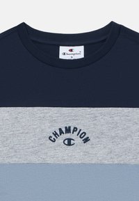 T-maica prugasta u tamnoplavoj i svijetlosivoj boji s okruglim izrezom. Sadrži "CHAMPION" logo u tamnoplavoj vezici na prsi. Pamuk.