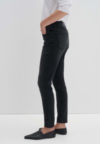 Jeans skinny noirs à taille haute, en denim texturé, avec cinq poches et une coupe ajustée. Portés avec des chaussures plates noires.