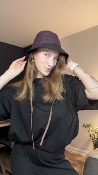 Chapéu tipo balde preto com uma faixa roxa escura, material leve, com uma tira para o queixo e textura suave. Usado com uma camisa preta larga.