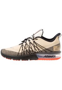 Baskets de sport beiges et grises avec une tige texturée, un système de laçage noir, un amorti à air transparent et une semelle extérieure orange, arborant des détails de marque.