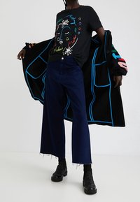 Svart t-shirt med färgstarkt ansiktsmotiv, kombinerad med mörkblå jeans med vida ben. En svart kofta med blå och rosa detaljer är slängd över. Svarta stövlar.