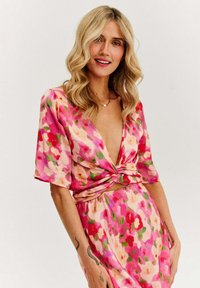 Květinové šaty v růžové barvě, s hlubokým výstřihem do V a krátkými rukávy. Materiál je lehký, s uzlovým detailem v pase a volným střihem.