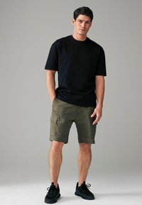 Zwarte t-shirt met korte mouwen, groene cargoshorts met zijzakken, en zwarte sneakers. Het model staat met de handen in de zakken tegen een neutrale achtergrond.