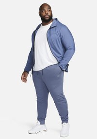 Lichtblauwe sportief jack met capuchon en ritssluiting, gecombineerd met bijpassende joggingsbroek. Witte sneakers maken de look compleet.