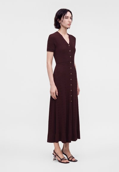 Femme debout dans une robe midi marron foncé boutonnée à manches courtes, portant des sandales noires à talons blocs avec brides.