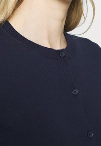 Námořnicky modrý cardigan z měkké pletené látky, s kulatým výstřihem a zapínáním na pět knoflíků ve stejné barvě.