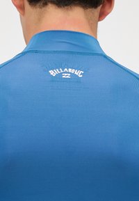 Zils izsargājošs krekls ar augstu apkakli, aizmugurē ar baltu "Billabong" logo, gluda tekstūra un pieguļošs dizains.