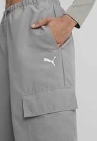 Svetlošedé cargo nohavice vyrobené z hladkej tkaniny, s bočnou vreckom a bielym logom Puma na stehne. Zahrnutý elastický pás.