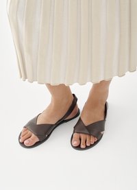 Kājām valkājot tumši brūnas ādas sandales ar krustenisku siksnu zem krēmkrāsas kroklota svārka uz balta grīdas.