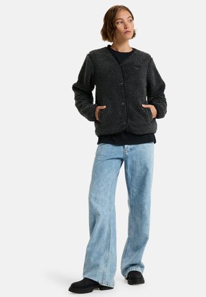 Zwarte fleecejas met een gestructureerd oppervlak, lange mouwen, knopen aan de voorkant en zijzakken, gecombineerd met lichte flared jeans en zwarte laarzen.