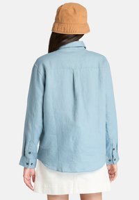 Lys blå, langermet skjorte laget av et strukturert stoff, med en bakfold og svarte knappdetaljer på ermene. Kombinert med en sandfarget bøttehatt.