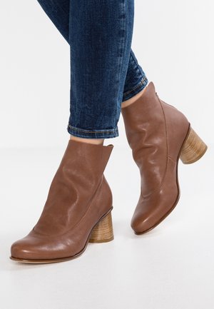 Bottines en cuir marron à talons blocs en bois portées avec un jean bleu foncé sur fond blanc.