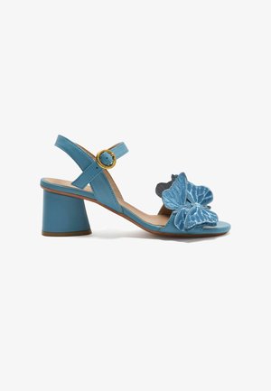 Sandalo in pelle blu con tacco basso, cinturino alla caviglia e fibbia dorata, con applicazioni decorative di farfalle blu sulla fascia frontale.