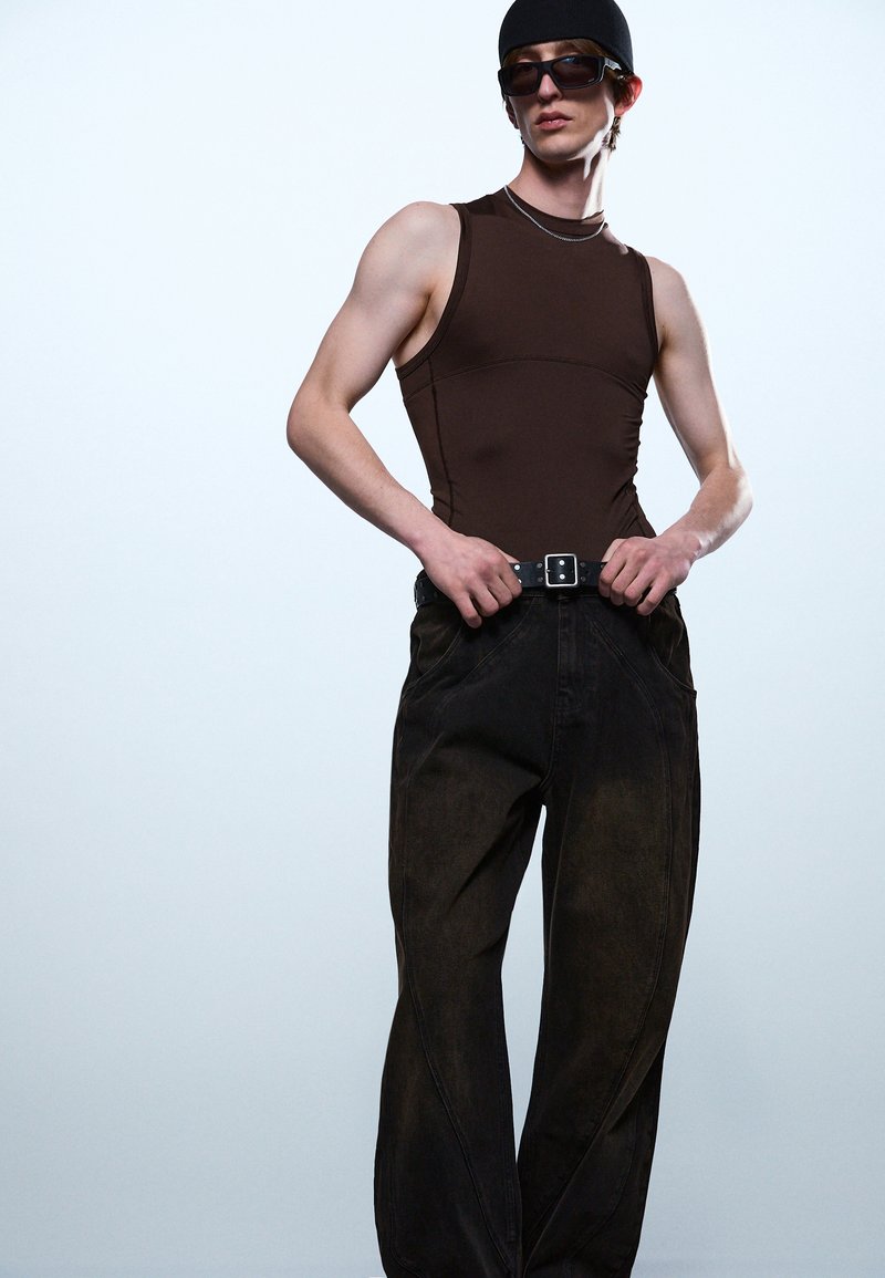 Tânăr purtând un top maro fără mâneci, pantaloni negri cu picior larg și curea, cagulă închisă la culoare și ochelari de soare, pe un fundal simplu deschis la culoare.