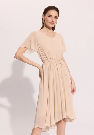 Kobieta w beżowej, zwiewnej sukience do kolan z krótkimi, falującymi rękawami, taliowaną w pasie i złotymi efektownymi kolczykami, pozująca z jedną ręką na biodrze.