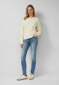 Crèmekleurige, gestructureerde trui met een ronde halslijn, gecombineerd met lichtblauwe skinny jeans en witte sneakers met een beige zool.