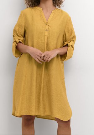 Femme portant une robe jaune moutarde longueur genou avec des manches retroussées et un col boutonné, debout devant un fond blanc uni.